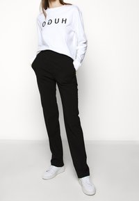 Sudadera blanca con la inscripción "HUGO" en letras negras, combinada con pantalones negros de corte tailored y zapatillas blancas, sobre un fondo liso.