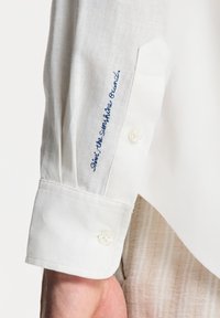 Weißes Langarmshirt aus strukturiertem Stoff. Verfügt über einen Knopfverschluss am Ärmel und blau gestickten Text am Ärmel. Einfaches Design mit sauberem Finish.