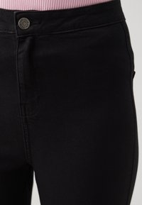 Jeans skinny de denim negro con cintura alta, que cuentan con un botón visible en la cinturilla y detalles de costura sutil a lo largo de las costuras.