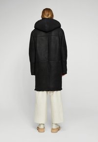 Abrigo de piel de oveja negra con capucha, con una silueta cuadrada y detalles de borde sin terminar, combinado con pantalones de pierna ancha de color claro y botas beige.