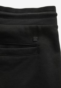 Joggers negros con una textura suave, que cuentan con una cinturilla elástica y un pequeño parche de logo cerca del bolsillo de un lado.