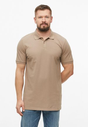 Homme barbu portant un polo beige à manches courtes et un jean bleu, debout devant un fond blanc uni.