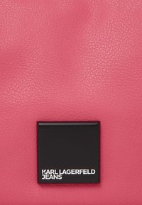 Ružičasta kožna torba s teksturiranom površinom, s crnom pravokutnom oznakom koja prikazuje "KARL LAGERFELD JEANS" u bijelom tekstu.