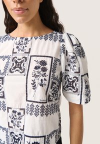Vit blus med puffärmar, prydd med intrikata marinblå blommönster och geometriska mönster, tillverkad av lättviktig bomullstyg.