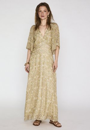 Femme portant une longue robe fleurie dorée avec des manches au coude, associée à des sandales marron et des bijoux en or, debout devant un fond blanc.