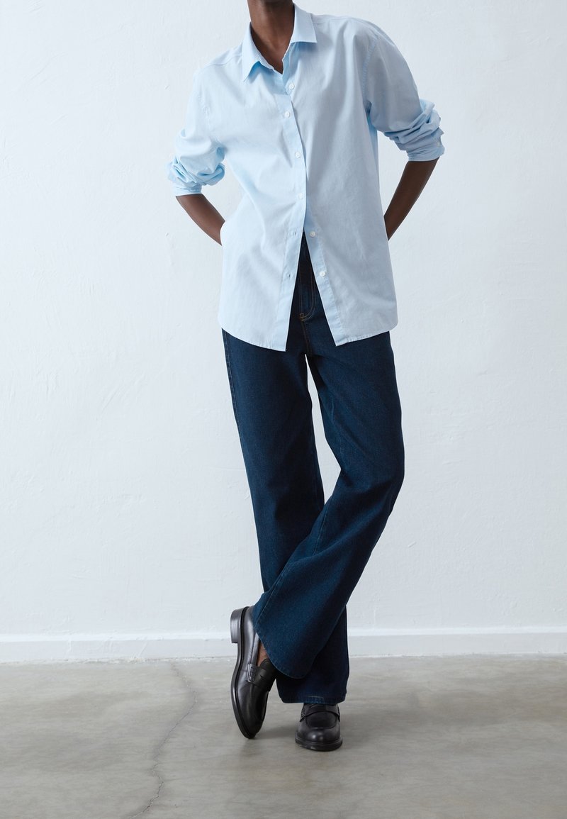 Chemise bleu clair à boutons avec manches retroussées, associée à un jean large bleu foncé et des chaussures noires, sur un fond uni.