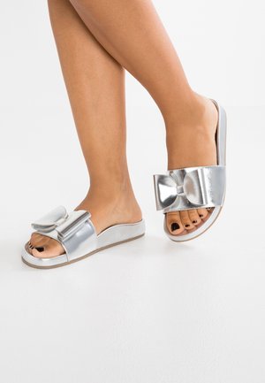 Srebrne klapki z dużym akcentem w postaci kokardy, otwarty przód, gładki metaliczny materiał i płaska podeszwa.