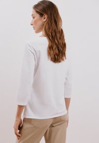Camicia bianca di cotone con maniche a tre quarti, scollo tondo semplice e vestibilità comoda, abbinata a pantaloni beige. Texture liscia.