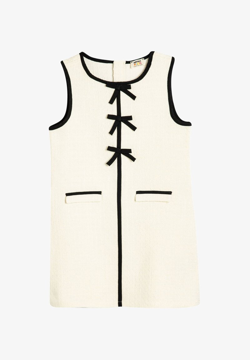 Valkoinen hihaton mekko, jossa on mustia yksityiskohtia ja kolme mustaa solmua etuosassa sekä kaksi etutaskua. Rakenteellinen kangas.