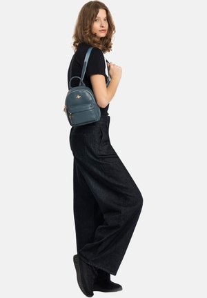 Giovane donna con capelli di media lunghezza indossa una t-shirt nera, pantaloni neri a gamba larga, sneakers nere e porta uno zainetto blu piccolo sulla spalla.