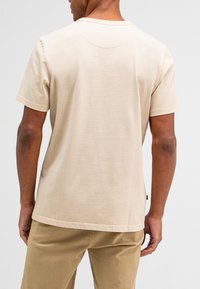 Beige T-shirt met korte mouwen en ronde hals, zachte textuur en een onderkant die iets rond is aan de achterkant. Casual pasvorm met minimale stiksels.