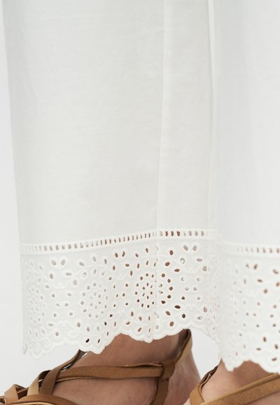 Λευκό ύφασμα με διακοσμητική δαντέλα στο τελείωμα, φορεμένο πάνω από πόδια σε καφέ δερμάτινα σανδάλια.