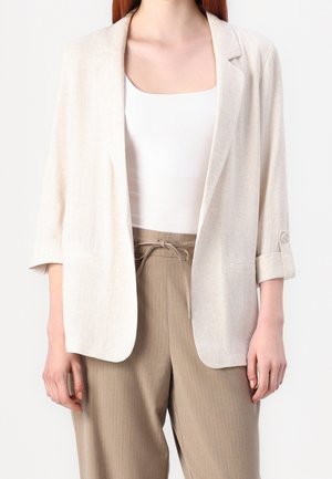 Femme portant un blazer beige clair à manches retroussées par-dessus un haut blanc et un pantalon beige à fines rayures taille haute noué à la taille.
