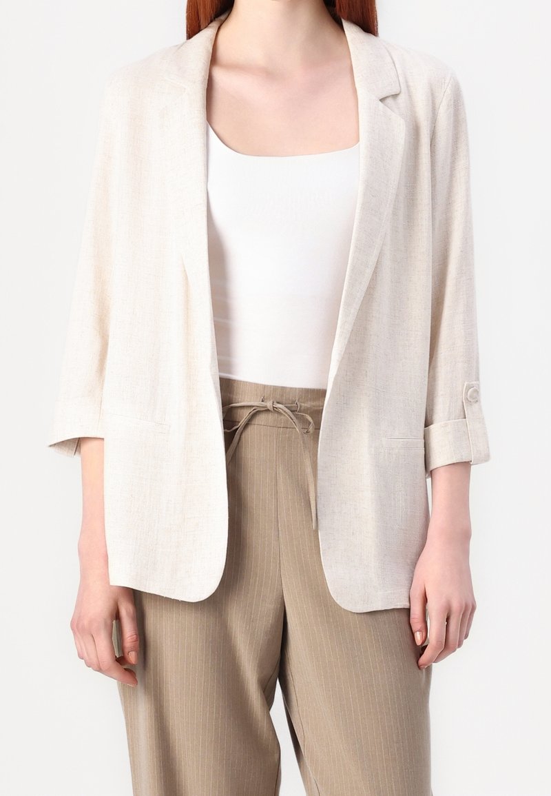 Femme portant un blazer beige clair à manches retroussées par-dessus un haut blanc et un pantalon beige à fines rayures taille haute noué à la taille.