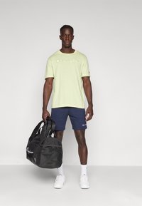 Light green t-shirt with "Champion" logo, navy shorts, white sneakers, and a black duffel bag with mesh detailing. Simple and modern design.