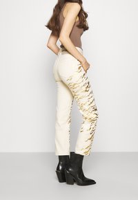 Jeans beige con patrón de rayas de tigre doradas, de cintura alta y corte recto, combinados con botas de tacón negro por encima del tobillo. Vista trasera, textura de tela suave.