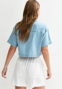 Camisa de mezclilla azul claro recortada con mangas cortas, que presenta una costura en el yugo posterior. Combinada con pantalones cortos beige claros con bolsillos traseros.