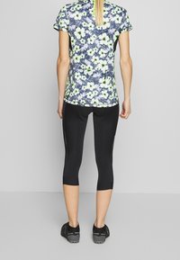 Kwiatowa koszulka sportowa z krótkimi rękawami w kolorze granatowym, białym i neonowozielonym, połączona z czarnymi legginsami do kostek i czarnymi butami sportowymi.