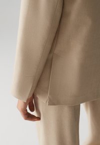 Beige op maat gemaakte blazer van een gestructureerde stof, met een splitdetail en een close-up van een hand die op de zijnaad rust.