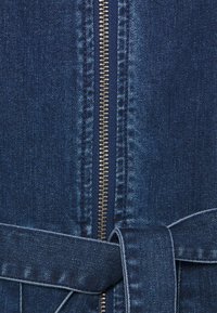 Gros plan sur un tissu en denim bleu foncé avec une fermeture éclair métallique centrale et une ceinture en denim nouée près du bas.