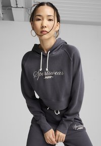 Tamno sivi dukserica s bijelim vezicama, na prednjoj strani ima izvezeno "Sportswear by PUMA". U kompletu su i odgovarajuće hlače sličnog brendinga.
