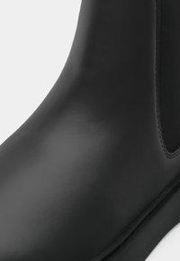Bota de borracha preta com uma textura lisa e brilhante, apresentando painéis laterais elásticos e um design de bico arredondado. A sola é plana e durável.