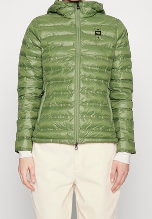 Henkilö, joka käyttää vetoketjullista, hupullista oliivinvihreää toppatakkia ja luonnonvalkoisia housuja, seisoo yksinkertaisen taustan edessä.