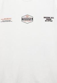 Biała bawełniana koszulka z logo Toyota Land Cruiser w kolorach czarnym i czerwonym. Na przodzie znajduje się napis "Driving All Roads in Style."