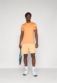 Oranžs sporta T-krekls ar melnu logotipu, apvienots ar gaiši oranžām šortiem. Figūra tur tenisam raketi un valkā baltas zeķes ar sporta apaviem.