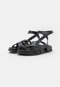 Schwarze Sandalen mit flacher Sohle, die überkreuzte Riemen und einen Knöchelriemen haben. Hergestellt aus einem glatten Material mit matter Oberfläche.