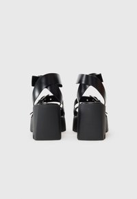 Sandalias negras de tacón con tacones rectangulares anchos. Presentan correas cruzadas y un acabado suave y brillante en el material superior.