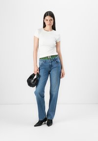 Hvit kortermet skjorte, blå denimjeans med grønt mønstret liv, og svart håndveske. Modell med svarte hælhæler.