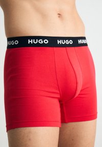 Sarkans kokvilnas bokseris ar melnu elastīgu jostu, kurā ir balti uzrakstīts "HUGO". Gluda tekstūra un pieguļošs dizains.