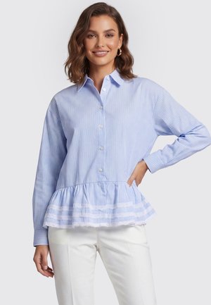 Woman wearing a blue and white striped button-up shirt with a ruffled hem and white pants, smiling with one hand on her hip.
