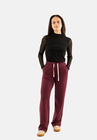 Haut noir à manches longues avec des manches transparentes, associé à un pantalon de survêtement bourgogne avec une taille élastique et un cordon blanc. Chaussures beiges.