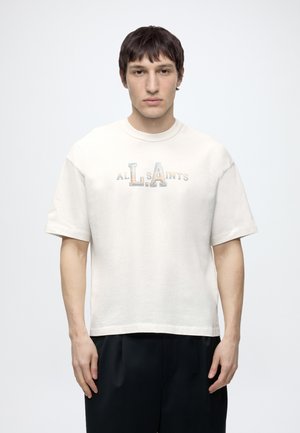 Mladý muž stojí čelom vpred, oblečený v bielej krátkoročnej tričku s nápisom „All Saints“ a čiernych nohaviciach na jednofarebnom pozadí.