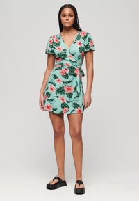 Mintgroene wikkeljurk met een bloemenpatroon met roze hibiscusbloemen en groene bladeren. Korte mouwen en een zijstrikdetail. Zwarte platform sandalen.