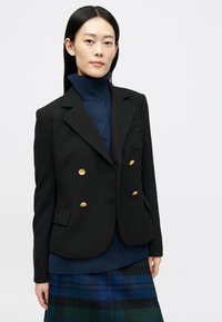 Črn dvoredni blazer z zlatimi gumbi, z teksturiranim pletenim ovratnikom. Obdan s temnomodrim puloverjem z visokim ovratom in karirasto krilom.