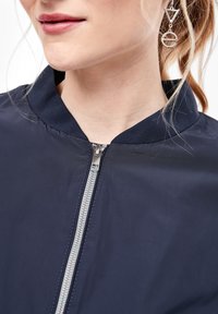 Femme portant une veste zippée bleu marine avec un col côtelé et une boucle d'oreille argentée géométrique en forme de triangle et de cercle.