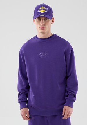 Młody mężczyzna w fioletowej czapce Los Angeles Lakers, bluzie i spodniach, stojący na jednolitym jasnym tle.