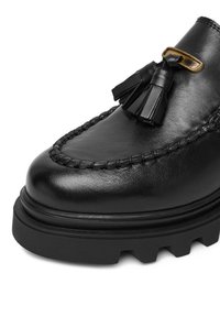 Čierny kožený mokasín s dekoráciou v podobe třásní, s pleteným švom pozdĺž okraja a hrubou gumovou podrážkou. Hladký povrch po celom diele.