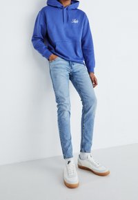 Person som har på sig en blå Levi's hoodie, ljusblå slim fit jeans och vita Nike sneakers med gummisulor, står mot en vit vägg.