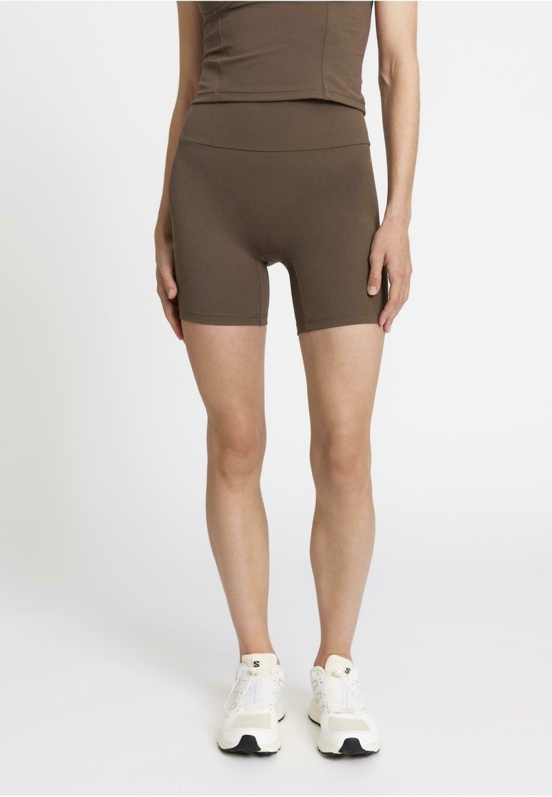 Pantalones cortos deportivos marrones con una cintura alta y una tela suave y elástica. Combinados con zapatillas blancas que tienen suelas negras y acentos de malla.