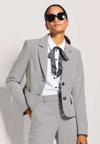 Grau strukturierter Anzug mit einem maßgeschneiderten Blazer und passenden Hosen. Weißes Hemd mit einer schwarzen Krawatte im Punkte-Design. Große schwarze Sonnenbrille.