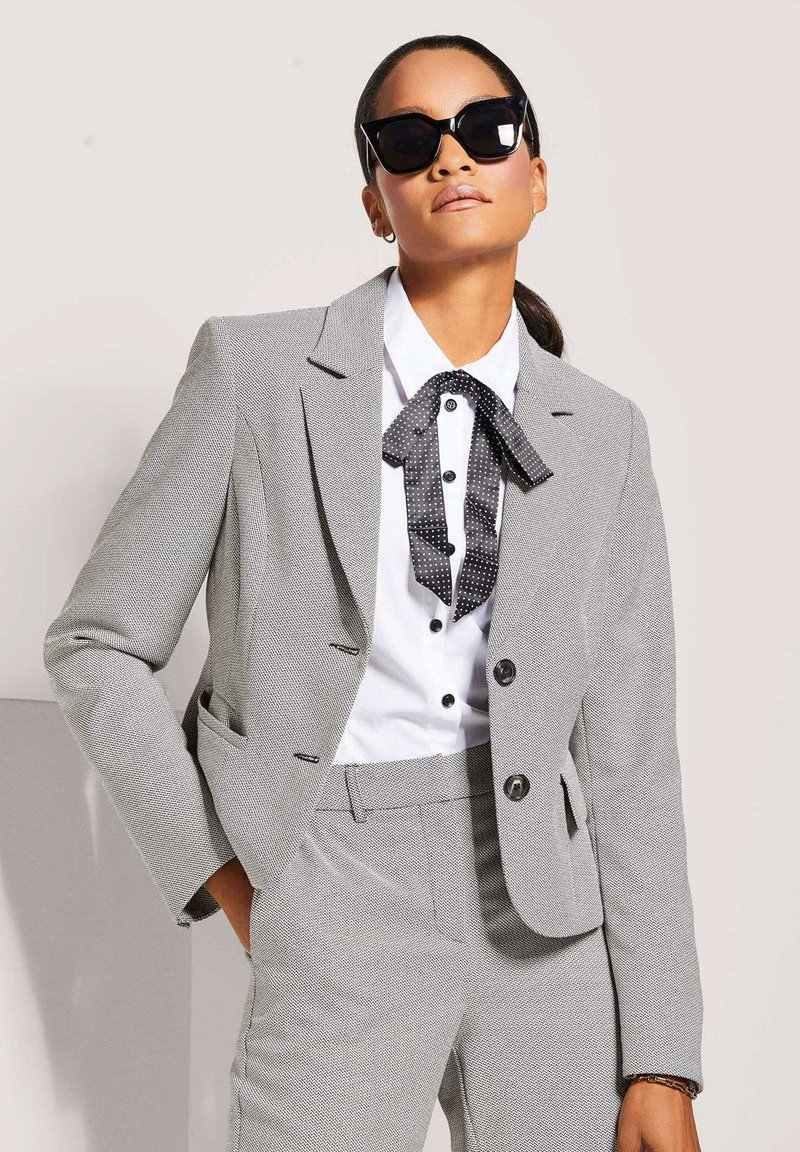 Grau strukturierter Anzug mit einem maßgeschneiderten Blazer und passenden Hosen. Weißes Hemd mit einer schwarzen Krawatte im Punkte-Design. Große schwarze Sonnenbrille.