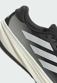 Baskets de sport noires avec une tige en maille texturée, comportant trois bandes blanches et une semelle intermédiaire blanche en couches. Les accents sur le talon sont noirs.