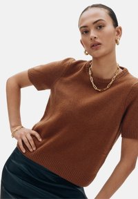 Pruun lühikeste varrukatega kampsun pehmest kangast, millel on sooniline kaelus ja äär, kombineeritud musta nahast seelikuga ja kuldsete kettidega aksessuaaridega.