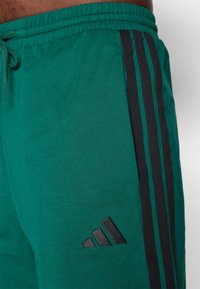 Grønne sweatpants med sorte tre-stripes detaljer, justerbar talje, og et broderet sort logo på siden. Blødt, bomuldsstof.