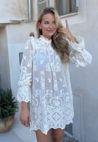 Robe blanche brodée avec des manches longues, décolleté volanté, motifs floraux et tissu transparent. Accentuée de bords de manches festonnés.