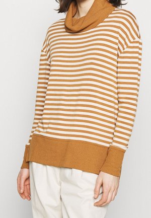 Frau trägt einen braun-weiß gestreiften Pullover mit Rippstrick-Rollkragen, Bündchen und Saum, kombiniert mit weißen Hosen.